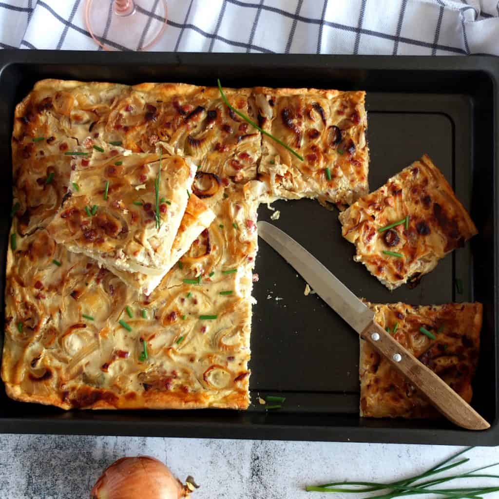 A black baking tray with onion cake. The onion cake is chopped up. you can see a knife with a wooden handle on the tray. Below the cake tray you can see an onion and some chives.