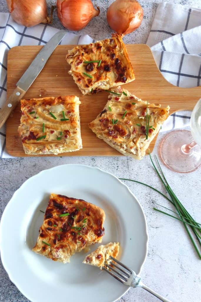 A white plate with a slice of onion cake on it. A piece is broken off with a fork. In the background you can see a chopping board with thee more slices of onion cake. On the top you can see some unpeeled onions