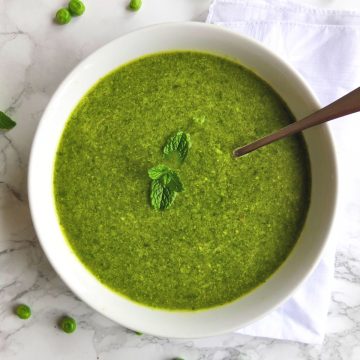 Pea soup in a white bowl. A mint leave on top. A spoon dunked in. Around you see single green peas