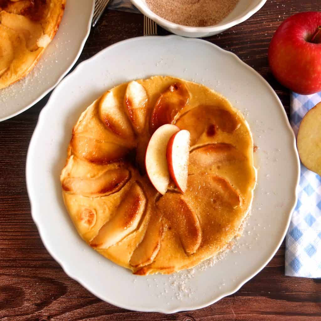 German apple pancake on a white plate that is set on a wooden background. On top of the pancakes there are two apple slices. Around the pancake there you can see a bowl of cinnamon sugar, an apple and part of a plate with another pancake