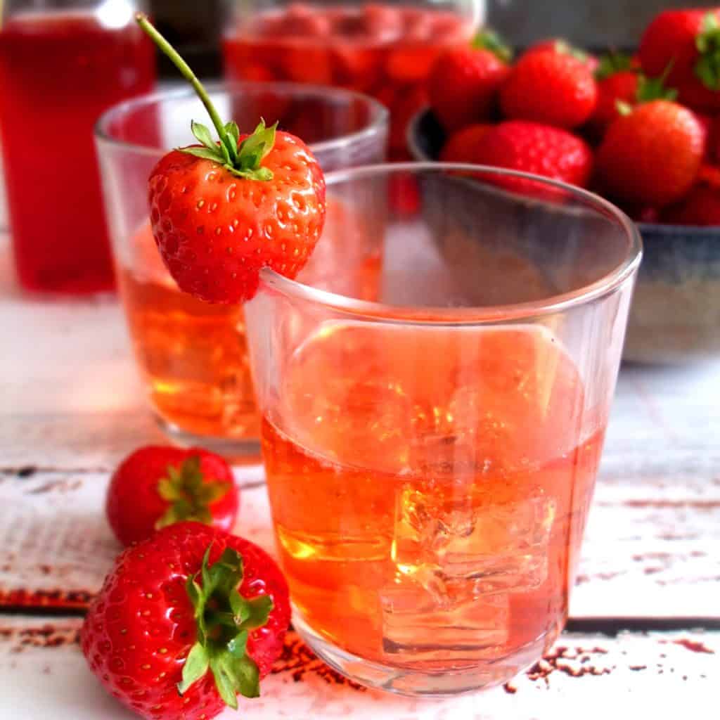 A glas with strawberry vodka lemonade on a white wooden background. On the rip of the glas is a strawberrz. In the background you can see a bowl of strawberries 