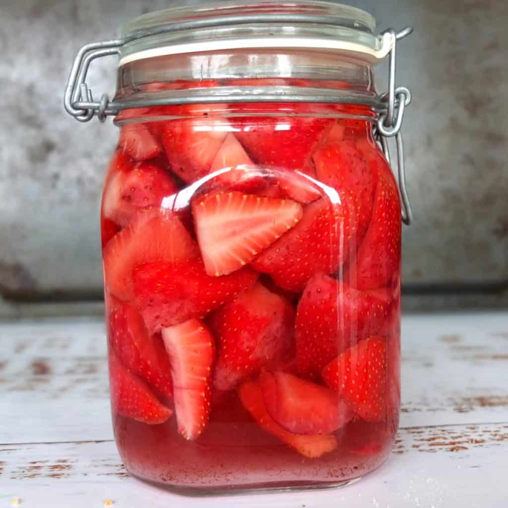 A mason jar on a white wooden surface. The background is grey. The jar is filled with red strawberries infused in vodka