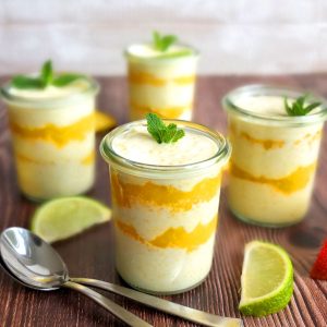 Four glasses filled with mango cream dessert. Each has a mint leave on top. Around you can see some limes and two spoons