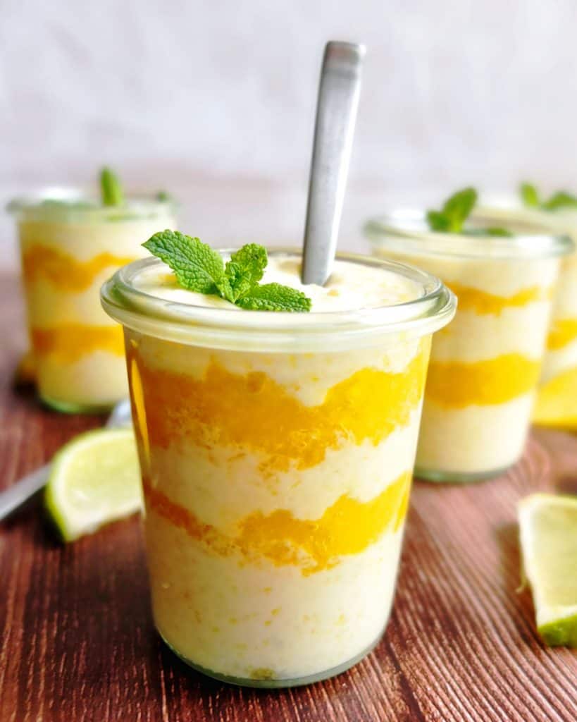 Layered mango and yoghurt dessert in a glass. A  spoon is sticking in the first glas. You can see three other glasses in the background. 