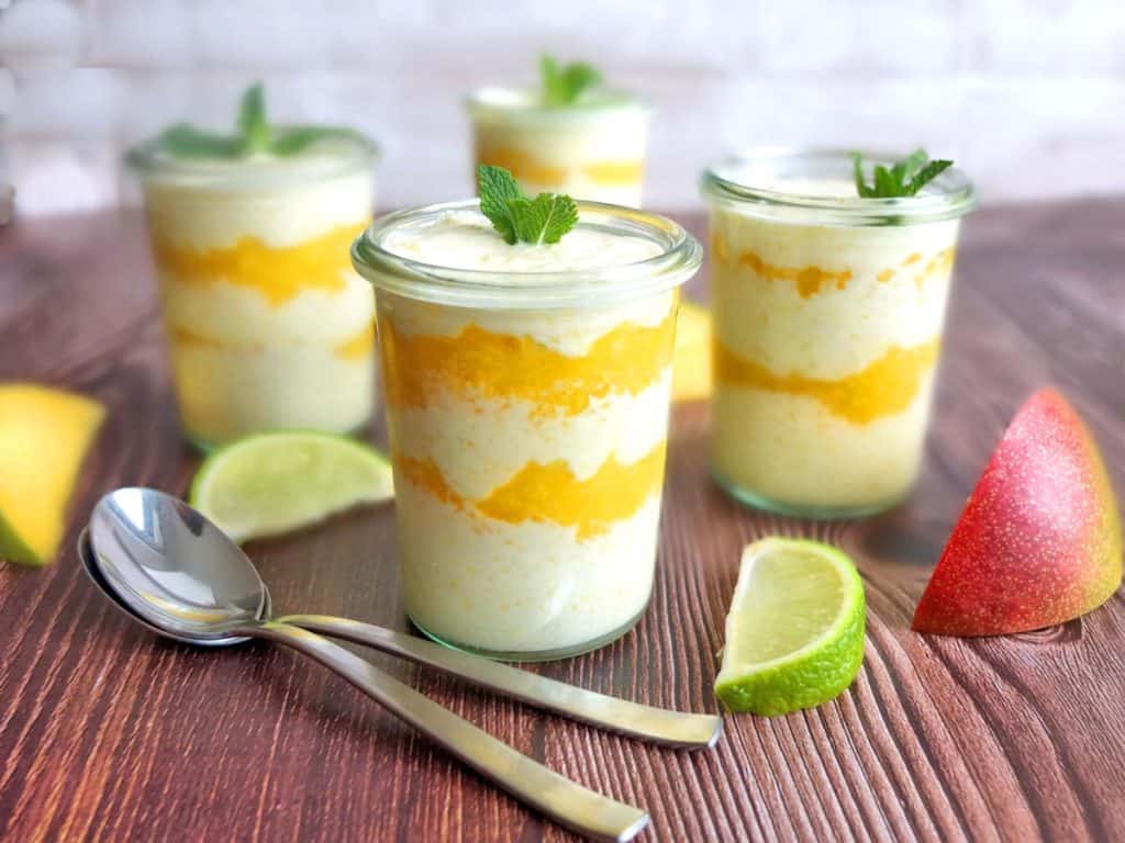 4 glasses filled with a layered mango dessert. Two spoons are at the front. Around you see some mangos and limes. 