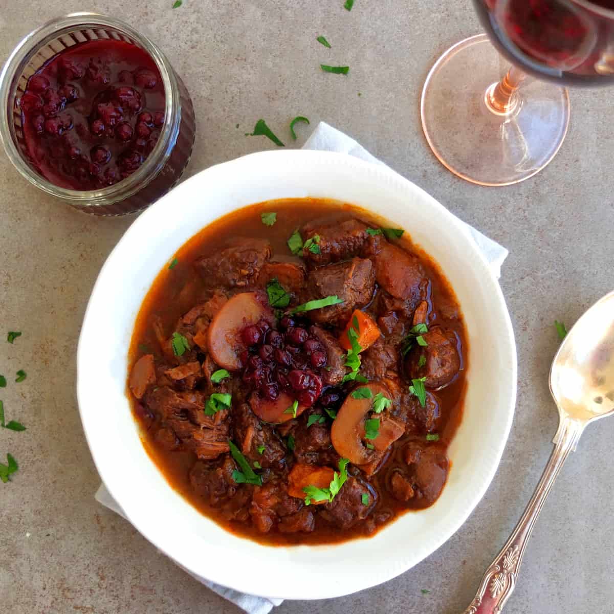A plate with venison goulash above a jar with cranberry sauce. You can see half a wine glass.
