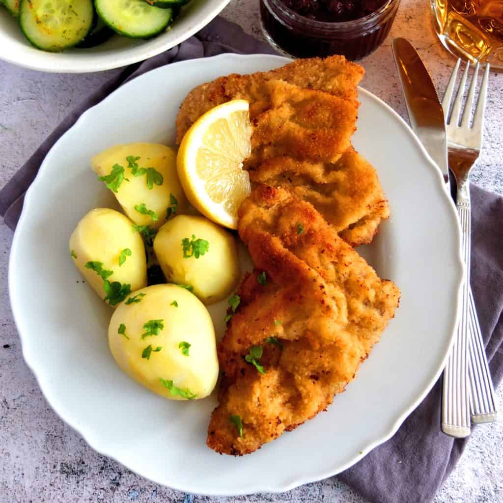 A Goldenbrown Vienna Schnitzel on a plate with parsley potatoes. At the top you can see a bowl of cucumber salad. The plate is white and on the right you can see a fork. 