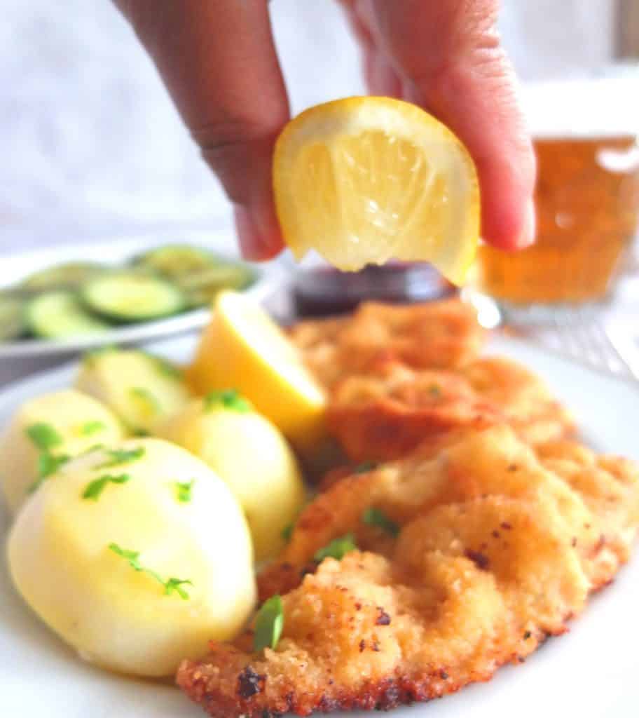 A Wiener Schnitzel on Parley potatoes on a white plate. Above you can see a lemon being squeezed over the