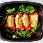 A black baking tray. On the baking tray there are some green lettuce leaves. On top of the lettuce leaves are potatoes and fish fillets The fish fillets in topped with breadcrumbs