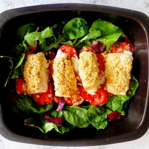 A black baking tray. On the baking tray there are some green lettuce leaves. On top of the lettuce leaves are potatoes and fish fillets The fish fillets in topped with breadcrumbs