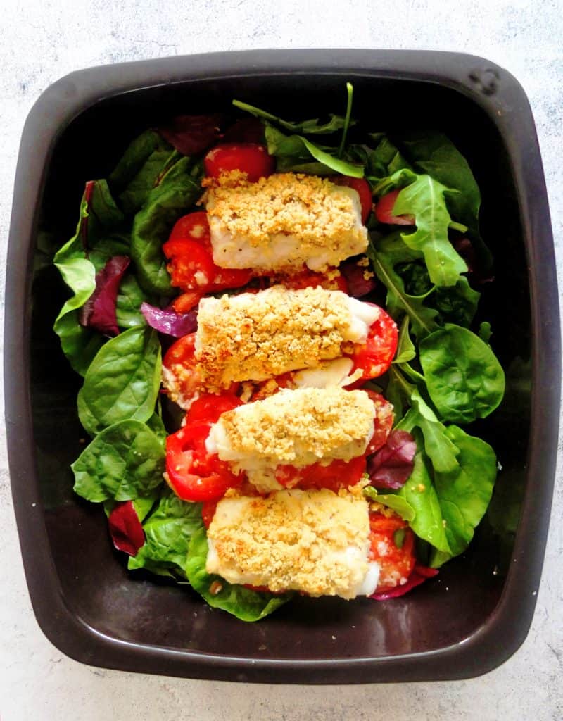 black baking tray. The baking tray is lined with green letuce leaves. In the middle there are some tomatoes and on top of the tomatoes there are some fish fillets. The fish fillets are topped with a parmesan crust.