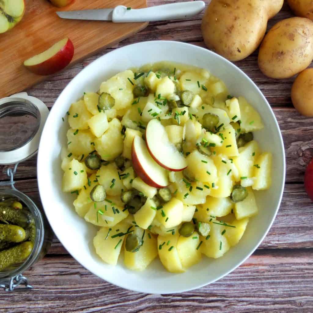 German Potato Salad on a White Plate. Around there is some cut up apple, a jar of Ghekins and some unpealed potatoes