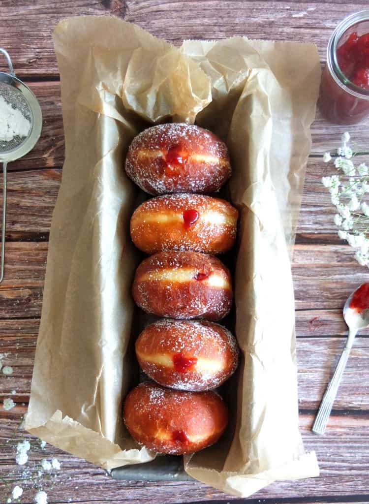 Berliner donut on top of some baking packment lined up in a tin. You can see some jam in the background and a spoon. 