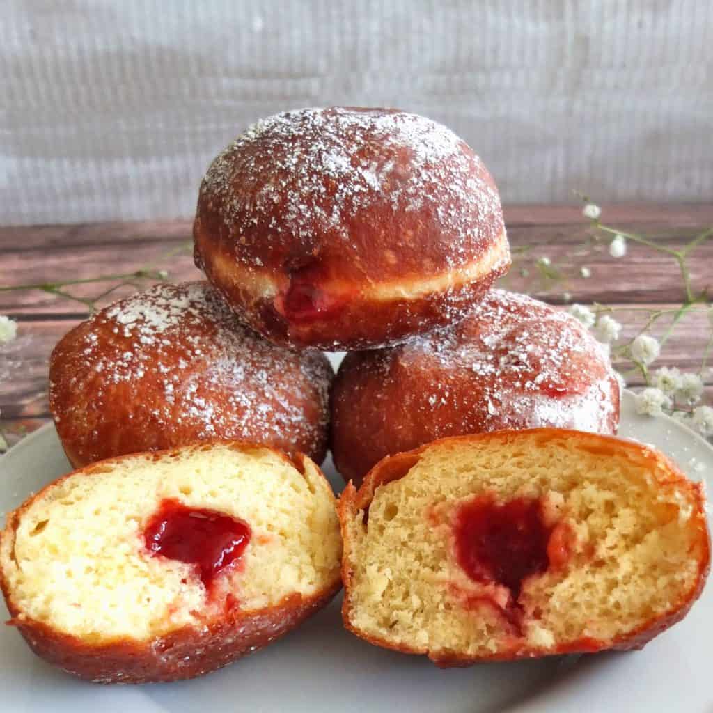 Four berliner donut on a plate. One is sliced in half and the jam is dripping out. The other three are stacked up as a pyramid