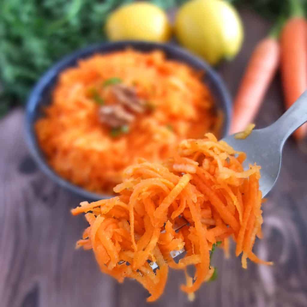 Carrot salad on a fork. In the background you can see the bowl of salad and some lemons and fresh carrots 