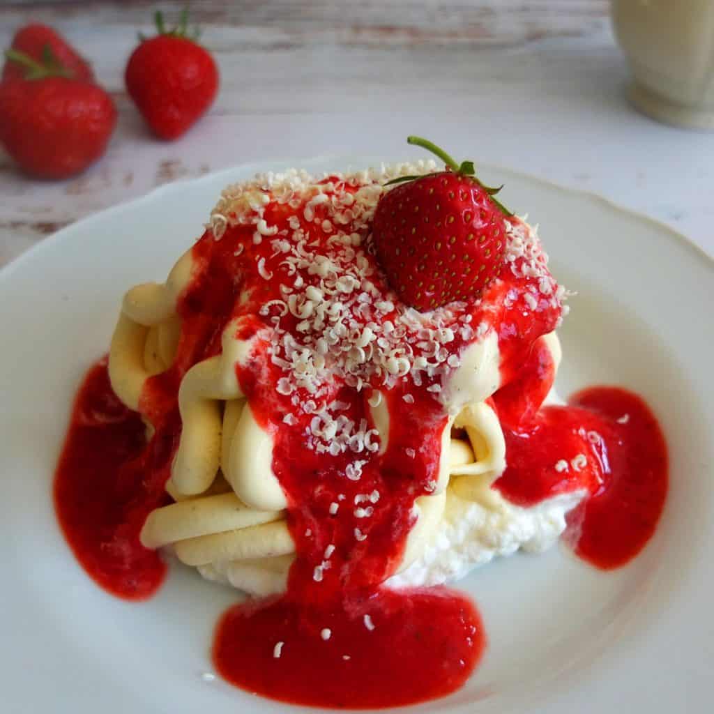 On a white plate, spagetti formed ice cream, with strawberry sauce and white chocolate sprinkles. On top a strawberry. In the background you can see further strawberries 