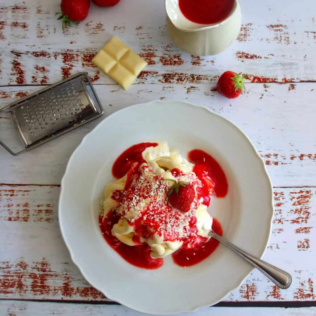 A plate with vanilla ice cream, topped with strawberry sauce. Above a saucier with strawberry icecream. Some fresh loose strawberries. A grater with white chocolate