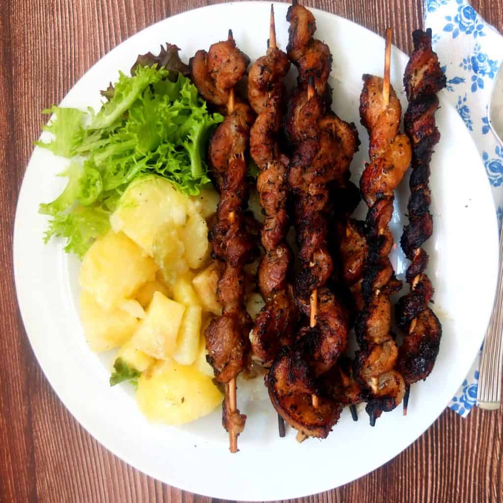 A white plate with Grillfackeln, bbqued pork belly on skewers. On the right you can see a portion of German potato sala and lettuce. On the left there is a blue white flowerd napkin. 