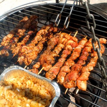 A bbq with pork belly skewers on the top. A tong is hovering of it. At the front you can see a aluminium bowl with onions