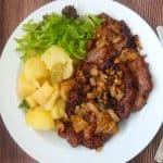 Schwenkbraten on a plate. Next to the meat is some german potato sala and lettuce. The Picture is on a wooden surface.