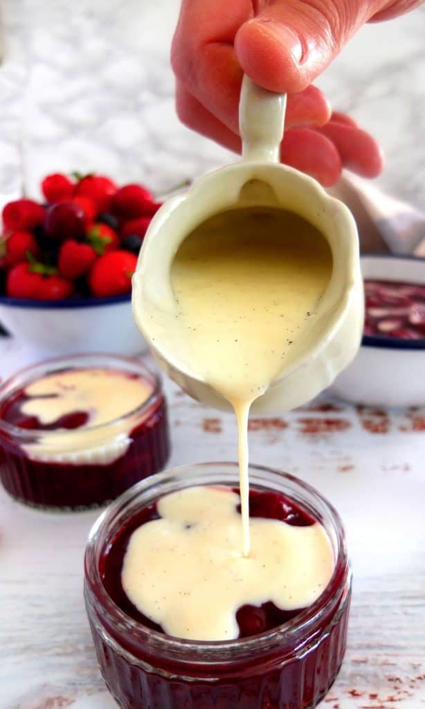 A hand holding a saucier of vanilla sauce. Pouring it on a glas bowl of Rote Gruetze. In the background another bowl of Rote Gruetze and a bowl of red berrie 