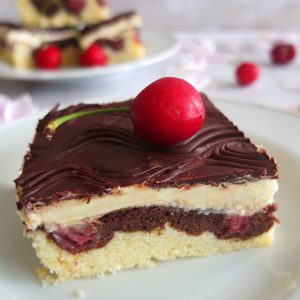 A slice of Danube Cake with a cherry on top. The slice of cake is topped by a cherry. In the background you can see a plate with more cake slices and some cherries.