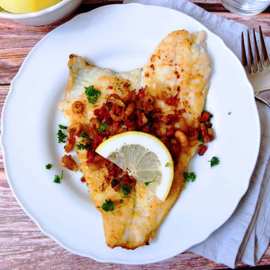 A white plate on a wooden background. On the plate you can see a fried plaice that is topped with bacon and brown shimp. A slice of lemon is on the fish. A fork is next to the plate. The dish is sprinkled with parsley.