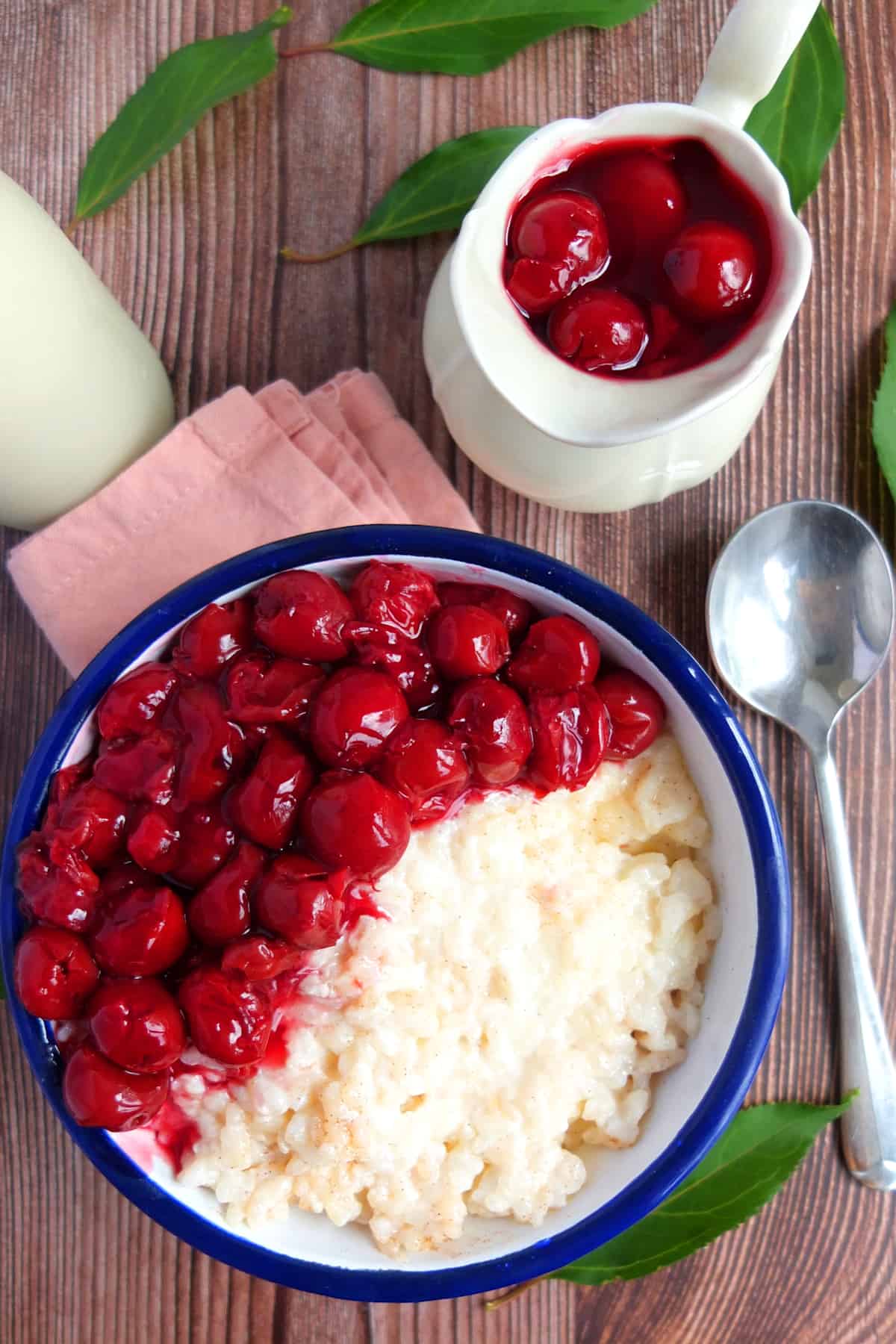 A white bowl with a blue rim. In it is German milchreis. Half of the rice is topped with some cherries. Above the bowl is a little dessert cream can with cherries. Next to the bowl is a spoon. The bowl sits on a pink napkin which is placed on a wooden background.