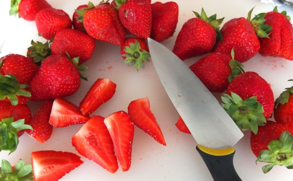 Strawberries on a white chopping board In the middle is a knife with a black handle. Some of the strawberries are chopped up. 