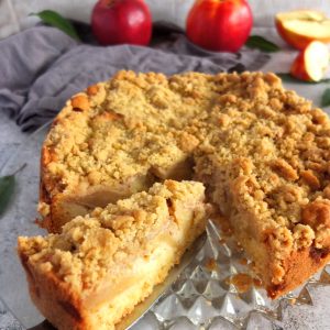 A German apple streusel cake on a glass plate. One of the slices is being lifted out with a cake slice. In the background there are some red apples. One red apple is sliced.