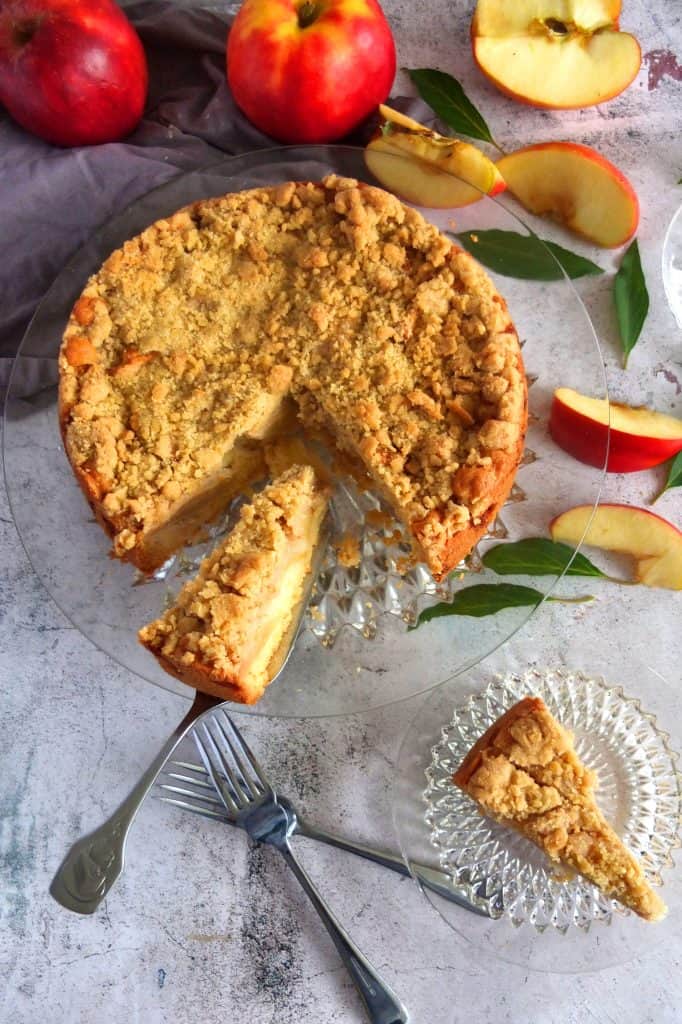 German apple cake on a glas platter. The cake plate is on a grew background. A piece of apple crumble cake is placed on a cake slicer. Lower down you can see two forks. A piece of apfelstreuselcake is on a glas plate. Above and around the plate are apples. Some of the apples are sliced.