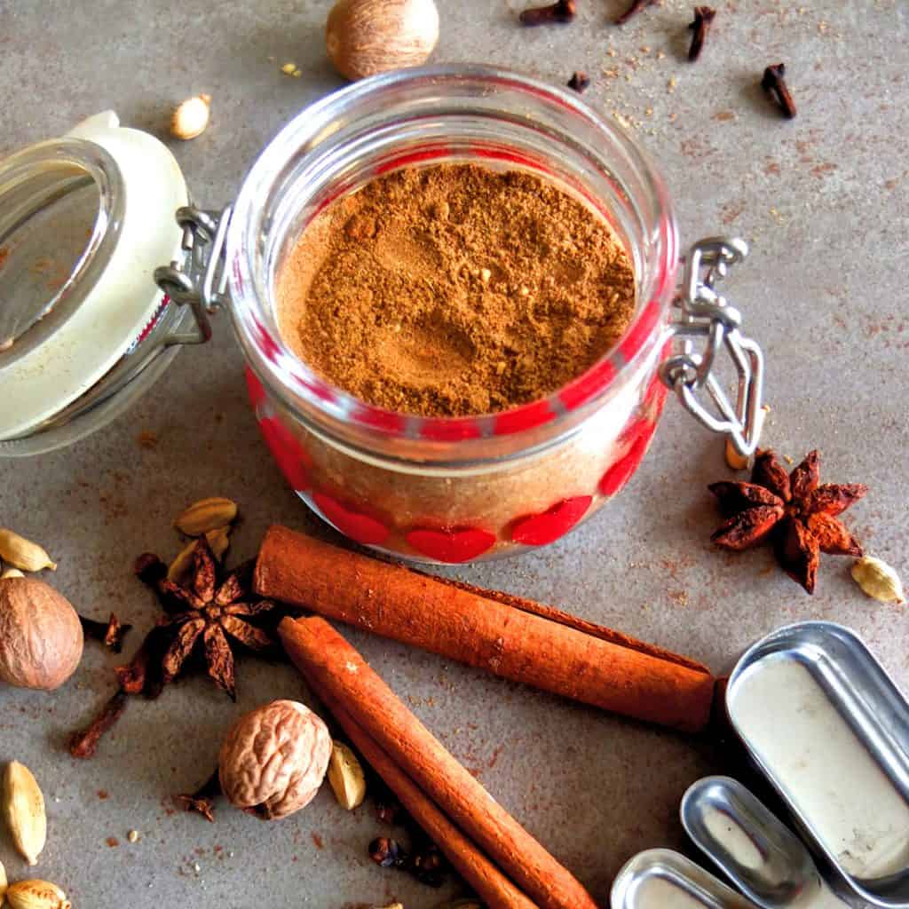 Lebkuchen Spice in a jar on a grez background. In front of the jar you can see cinnamon sticks, nutmegs, star anice. In the background there are some cloves. In the right corner you can see some measuring spoons