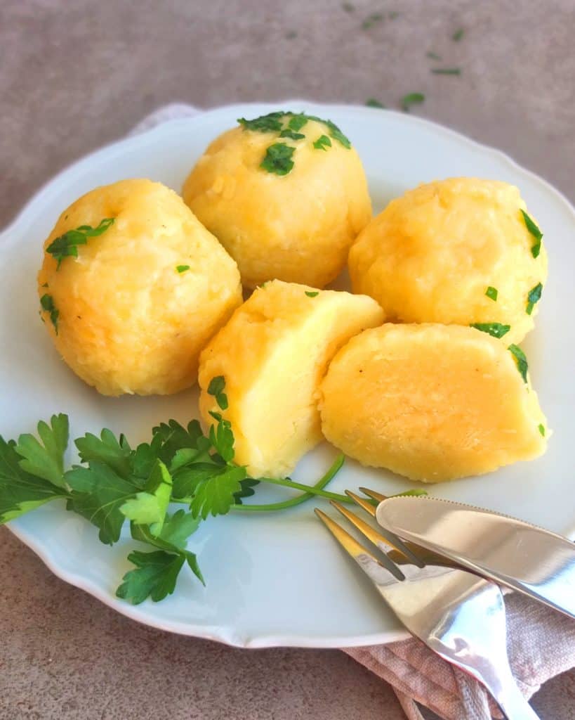 German Potato Dumplings on a white plate sprinkled with parsley butter. One Dumpling is cut in half. There is a fork and knife resting on the plate