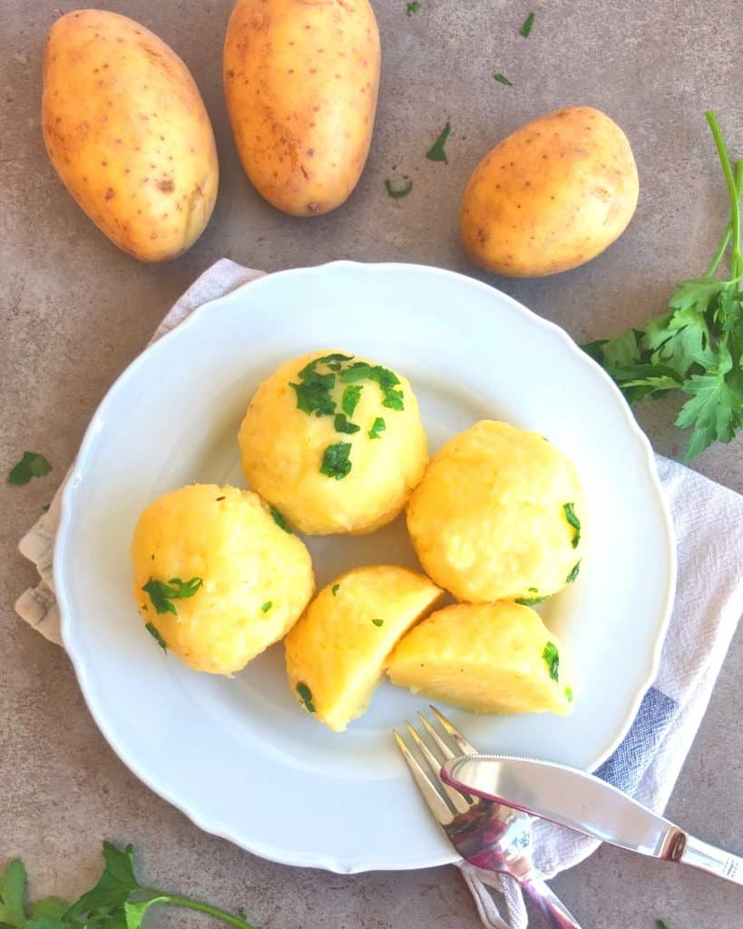 Kartoffelknödel on a  white play set on a grey background. The Knödel are sprinkled with parsley butter. Above the plate you see some raw potatoes and on the left a bunch of parsley. A fork and knife are resting on the plate