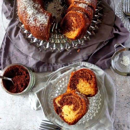 German Marble Bundt Cake. Two pieces on a glas plate. In the background you can see the bundt cake already cut. On the left is a pot of cocoa