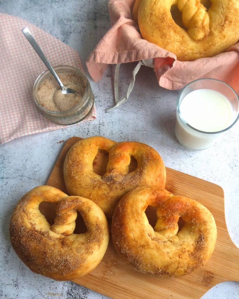 German Cinnamon Sugar Pretzels on a chopping board. 5 layed out on a white surface. A bowl of cinnamon sugar next to it. A basked with more pretzels. A Glas of milk is next to it.