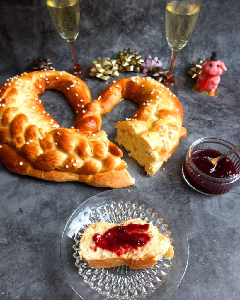 New Years pretzel on grey background. In the background you can see two champagne glasses and a marzipan pig and some golden ribbons. A slice is cut off and in the foreground is a slice of yeast bread with jam