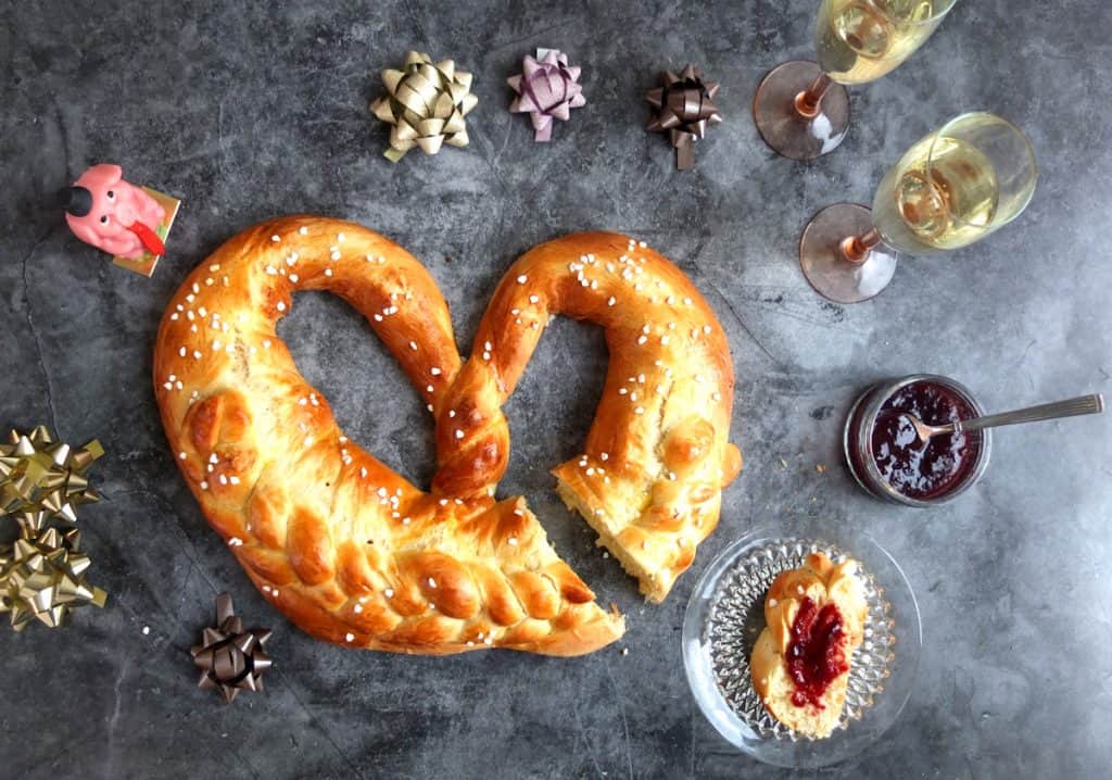 New Years pretzel on grey background. Around the pretzel you can see two champagne glasses and a marzipan pig and some golden ribbons. A slice is cut off and in the foreground is a slice of yeast bread with jam