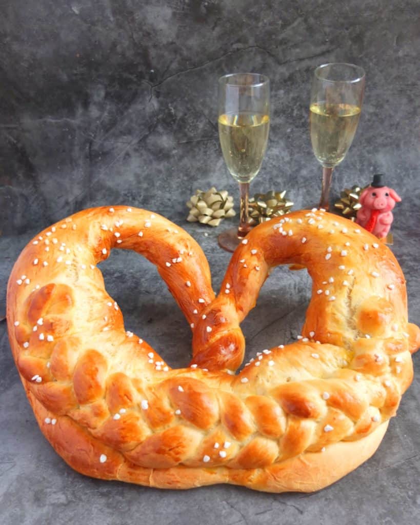 New Years pretzel on grey background. In the background you can see two champagne glasses and a marzipan pig and some golden ribbons