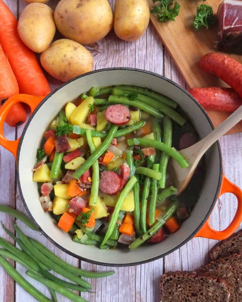 A saucepan with green bean soup. Around you can see the ingredients such as the sausages, bacon, carrots, apples, beans and bread