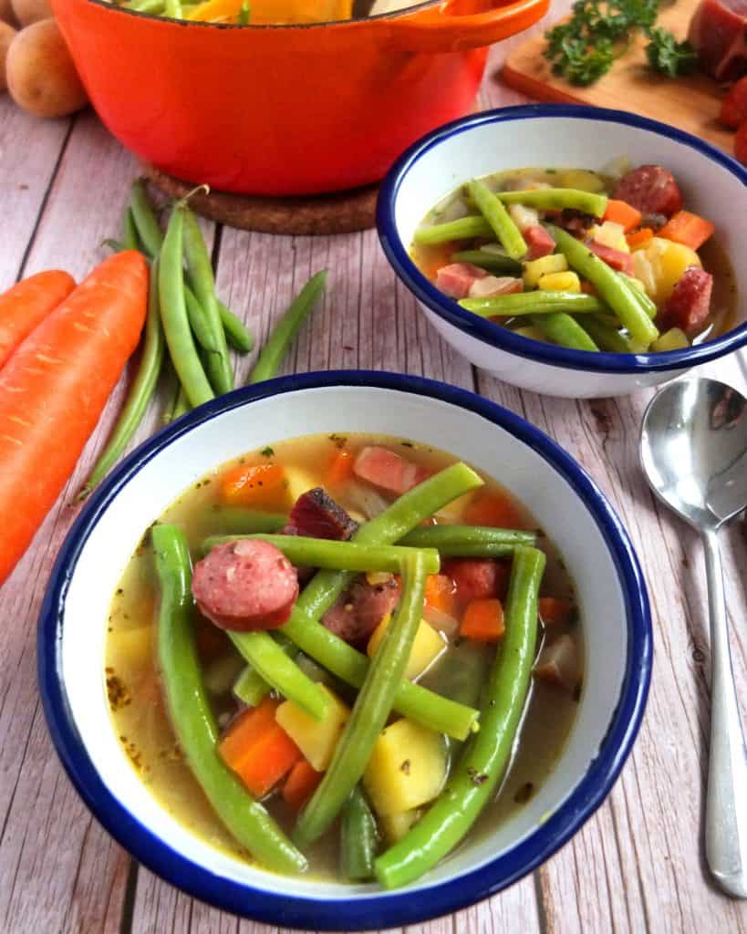 White and Blue bowl of Green Bean soup. In the background a saucepan and a further bowl of soup. Around you can see the ingrdients such as green bean, carrots and potatoes
