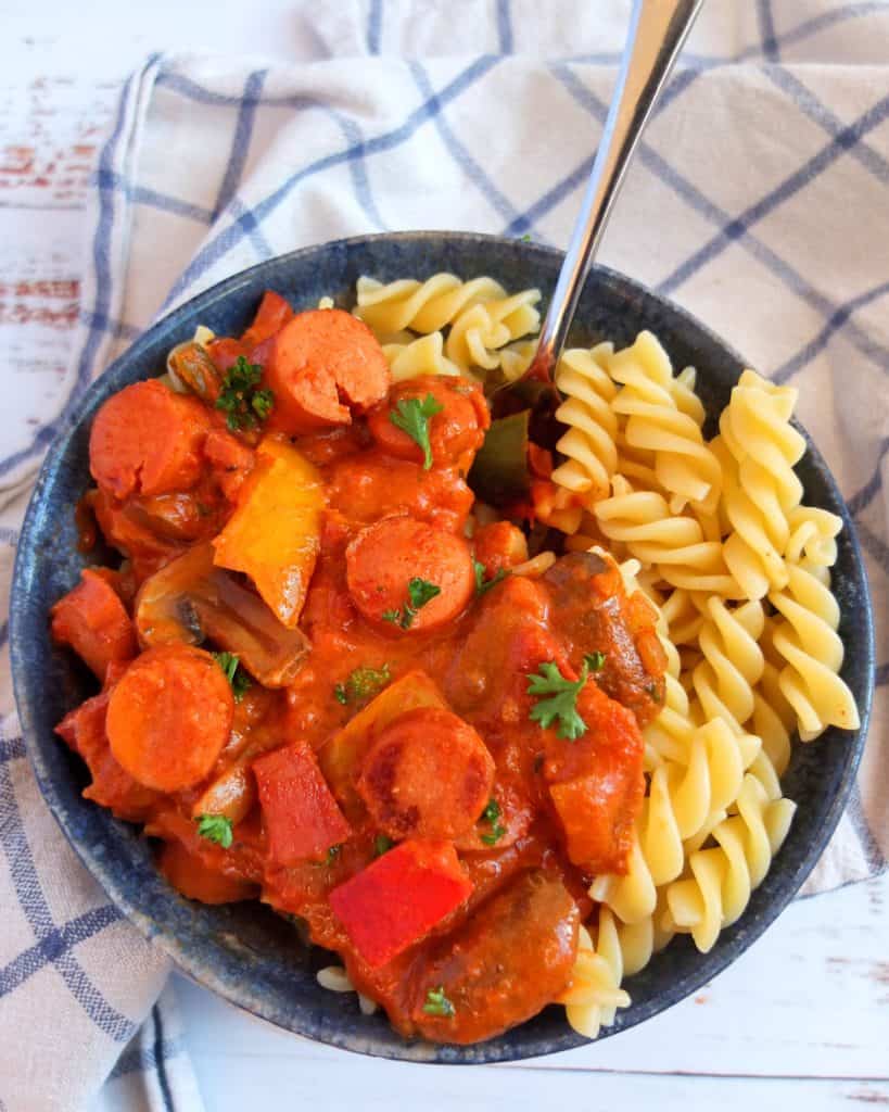 a bowl of sausage goulash with pasta on a blue and white checked teatowel.