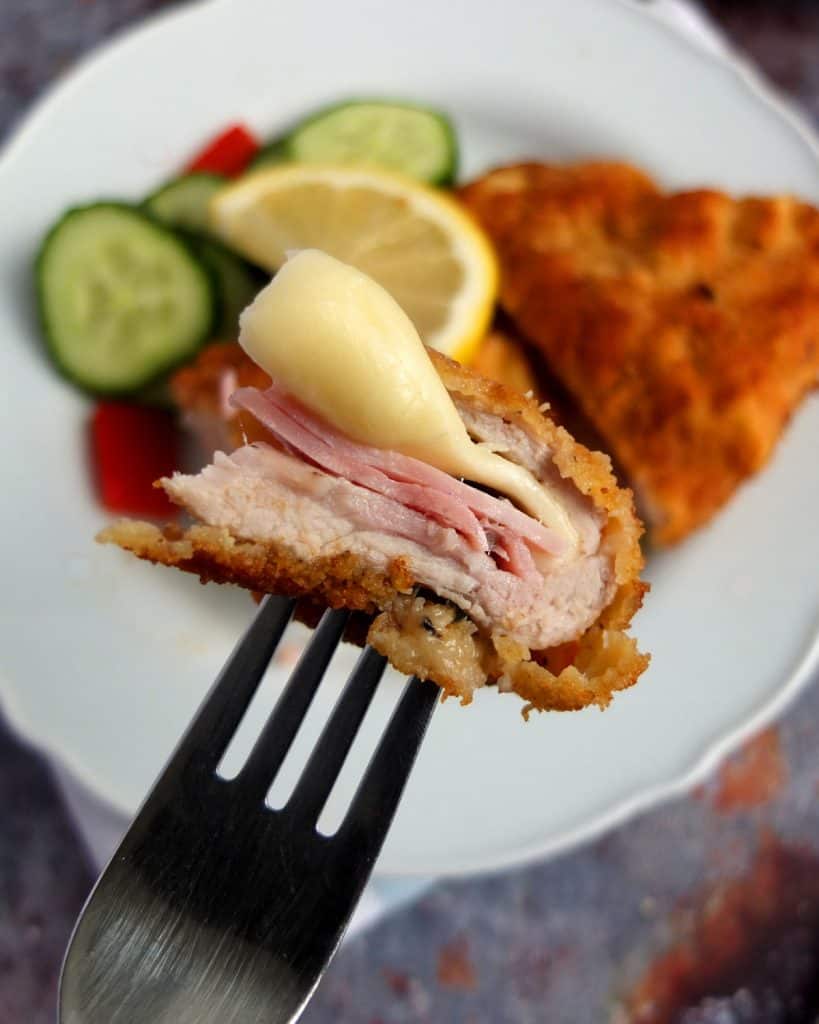 A piece of pork cordon bleu on a fork. In the background a plate with the rest of the schnitzel.